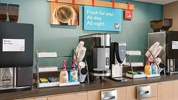 Una estación de café con varias máquinas de café, un letrero que dice “Recién hecho para ti. Todo el día. Toda la noche.” y un molinillo de café de pared.