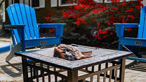 Zwei blaue Adirondack-Stühle mit einem Tisch dazwischen, ein Feuerkorb und ein Backsteinhaus im Hintergrund mit roten Blättern.