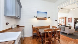 Un comedor con mesa y sillas de madera, pared blanca con un cuadro, y área de cocina con armarios de madera y un lavamanos blanco.