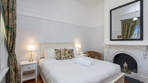 Un dormitorio con cama, dos mesitas de noche con lámparas, una silla, chimenea y un espejo en la pared.