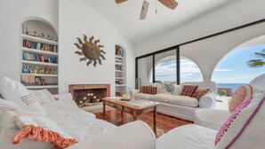 A spacious living room with white furniture, a fireplace, bookshelves, and a large window with a view of the ocean. - Casa Alvarado | Beachfront Private Escape w/ Pool (Baja California Sur)