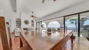 A spacious living area with a large wooden table, white sofas, and a bookshelf. Large windows offer a view of the ocean and palm trees. - Casa Alvarado | Beachfront Private Escape w/ Pool (Baja California Sur)