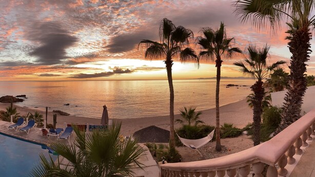 A poolside view with lounge chairs, a hammock, and palm trees overlooking a beach at sunset. - Casa Alvarado | Beachfront Private Escape w/ Pool (Baja California Sur)