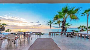 An outdoor seating area with chairs and a table, overlooking a beach with palm trees and a sunset. - Casa Alvarado | Beachfront Private Escape w/ Pool (Baja California Sur)