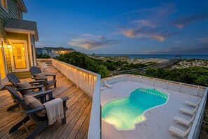 A wooden deck with a swimming pool, lounge chairs, and a view of the beach and ocean.