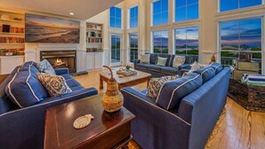 A spacious living room with a fireplace, large windows, and a view of the outdoors. - Oceanfront Estate | 14BR | Private Pool & Hot Tub | Sleeps 32 (Corolla)