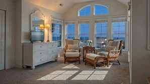 A spacious room with a large window, a television, a white dresser, and two wicker chairs. - Oceanfront Estate | 14BR | Private Pool & Hot Tub | Sleeps 32 (Corolla)