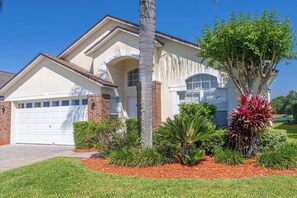 A two-story house with a garage, surrounded by a well-maintained garden with various plants and a palm tree. - Tropical Boho Lakefront Villa | Private Pool & Spa | Game Room | Near Disney (Kissimmee)