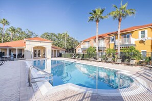 A swimming pool with a sunbathing area, surrounded by palm trees and buildings with red roofs. - Tropical Boho Lakefront Villa | Private Pool & Spa | Game Room | Near Disney (Kissimmee)