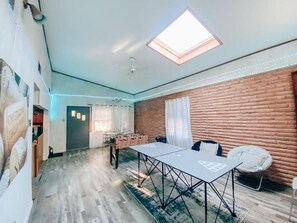 A room with a wooden floor, a table with a logo, a foosball table, a round chair, a window with curtains, and a skylight. - Middle of Dallas/Fort Worth! WORLD SERIES, RANGERS, COWBOYS, MAVS, CONCERTS!! (Irving)
