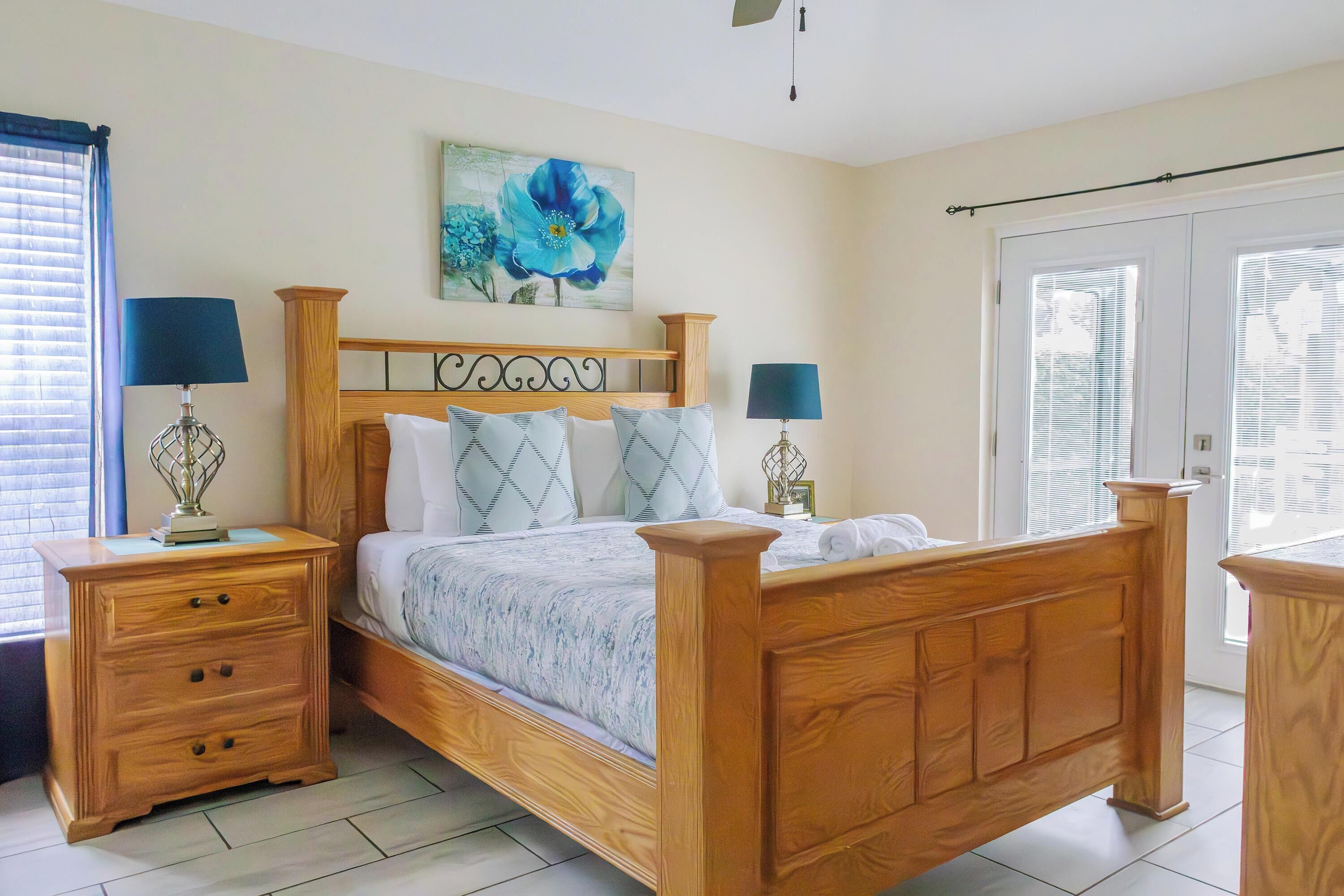A bedroom with a wooden bed, two nightstands with lamps, a large window with blinds, and a painting of flowers on the wall.