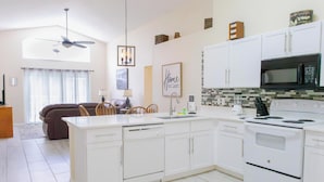A modern kitchen with white cabinetry, a built-in microwave, and a stove. There is a dining area with a brown sofa and wooden chairs.