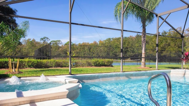 A swimming pool with a hot tub, surrounded by a covered area with a view of trees and a grassy area.