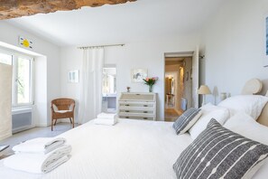 Un dormitorio con cama, cómoda, silla y baño visible a través de una puerta abierta.