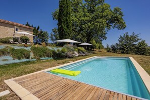 A rectangular swimming pool with a wooden deck, surrounded by lounge chairs and umbrellas, with a stone building and trees in the background. - Stone Cabin with Swimming Pool - King-Size Beds & Views of the Pyrenees (Gondrin)