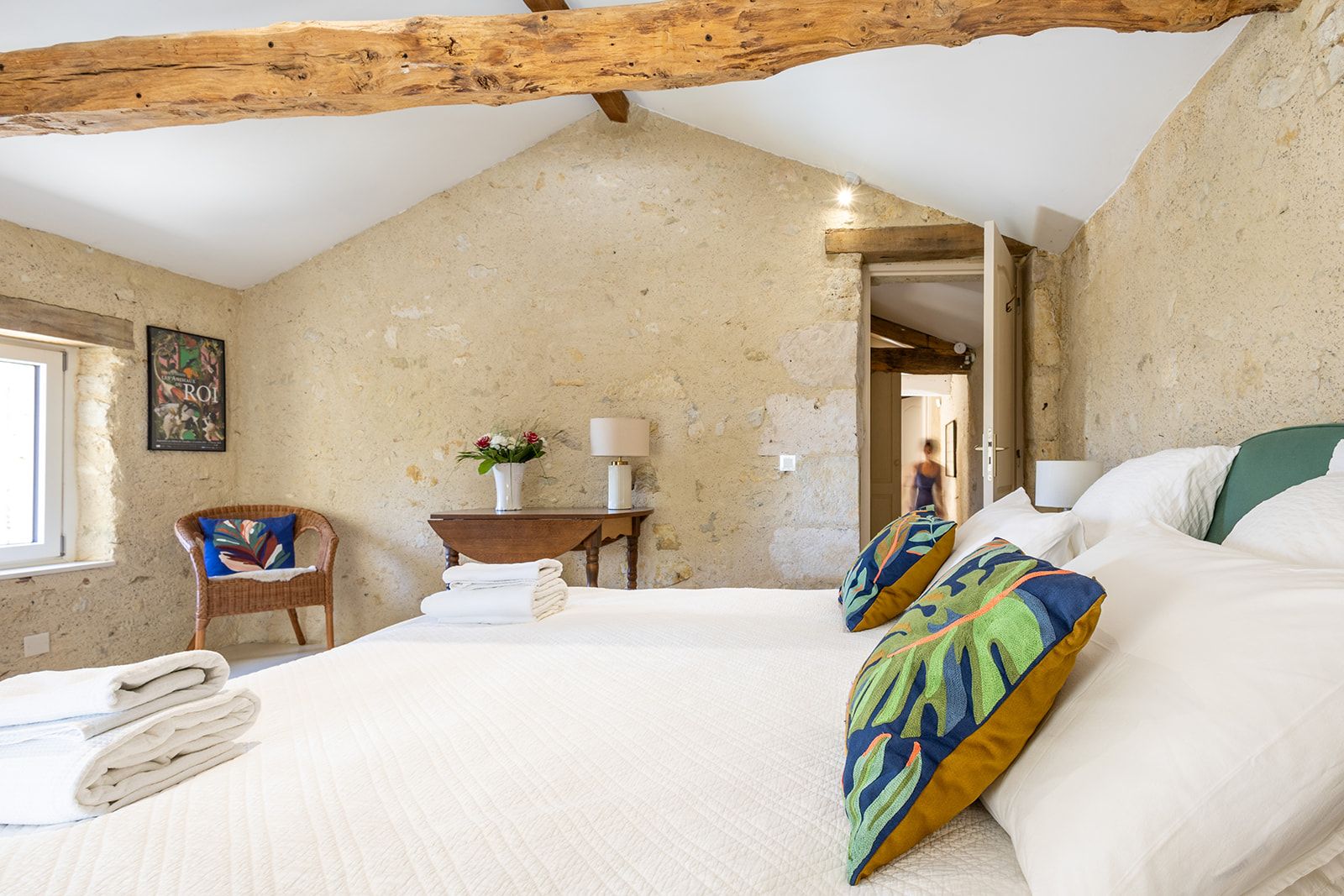 Een slaapkamer met een schuine kap, een bed met witte lakens, een stoel met een kleurrijk kussen, een nachtkastje met een lamp en een aan de muur hangend schilderij.