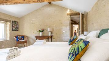 Een slaapkamer met een schuine kap, een bed met witte lakens, een stoel met een kleurrijk kussen, een nachtkastje met een lamp en een aan de muur hangend schilderij.
