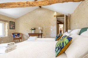 A bedroom with a sloped ceiling, a bed with white linens, a chair with a colorful cushion, a bedside table with a lamp, and a wall-mounted picture. - Stone Cabin with Swimming Pool - King-Size Beds & Views of the Pyrenees (Gondrin)