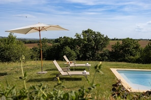 A poolside area with two lounge chairs and a large umbrella, surrounded by lush greenery and a view of rolling hills.