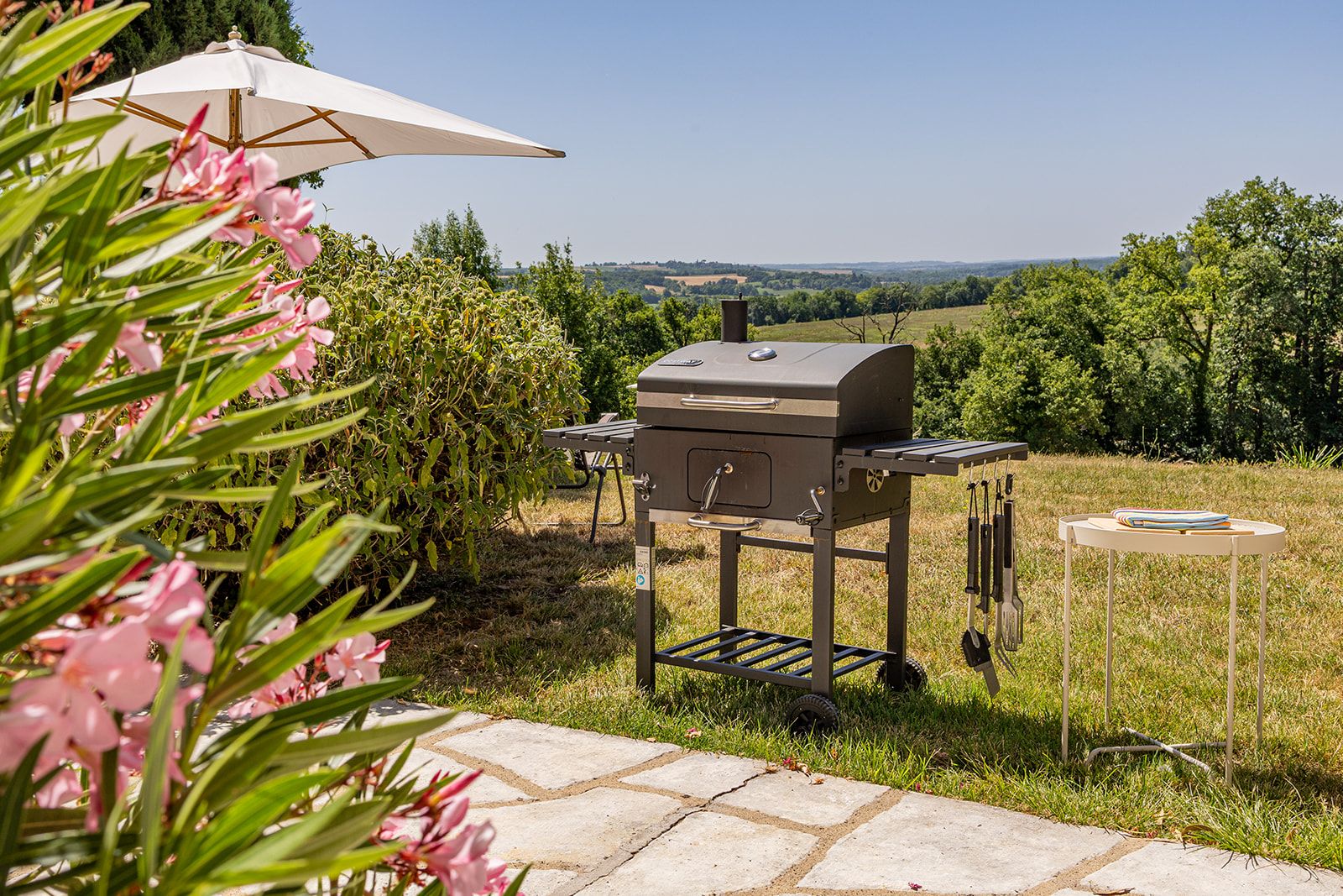 An outdoor grill setup with a table and tools, surrounded by greenery and a clear view of the landscape.