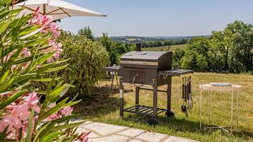 An outdoor grill setup with a table and tools, surrounded by greenery and a clear view of the landscape.
