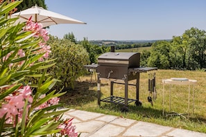 Un asador al aire libre con mesa y utensilios, rodeado de vegetación y una vista despejada del paisaje.