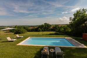 A rectangular pool with a wooden deck, surrounded by a grassy lawn and trees. There are lounge chairs and a table with a parasol.