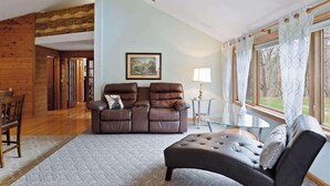 A cozy living room with a brown leather sofa, a glass coffee table, and a large window with curtains.