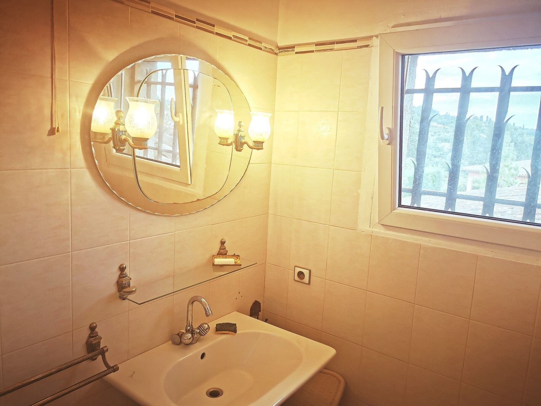 Een badkamer met een ronde spiegel, een wastafel en een raam met tralies.
