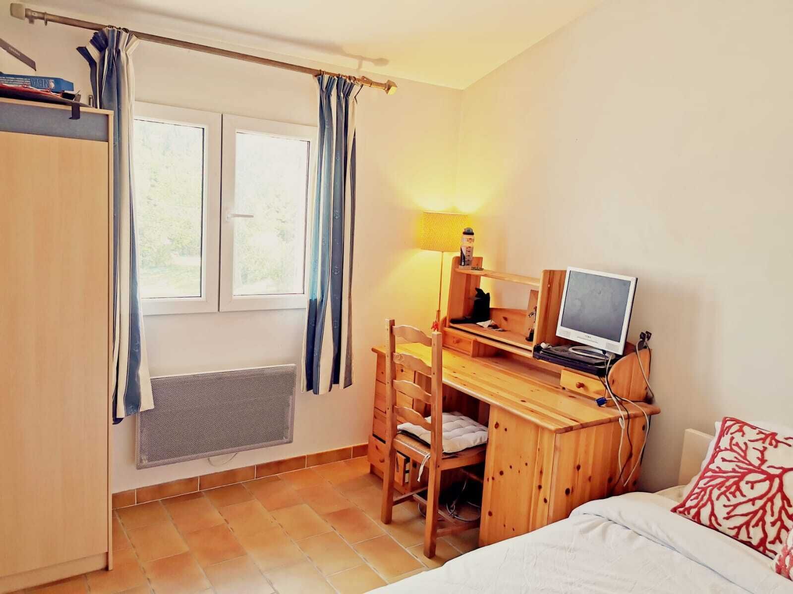 A compact bedroom with a bed, a desk with a computer, a chair, and a window with curtains.
