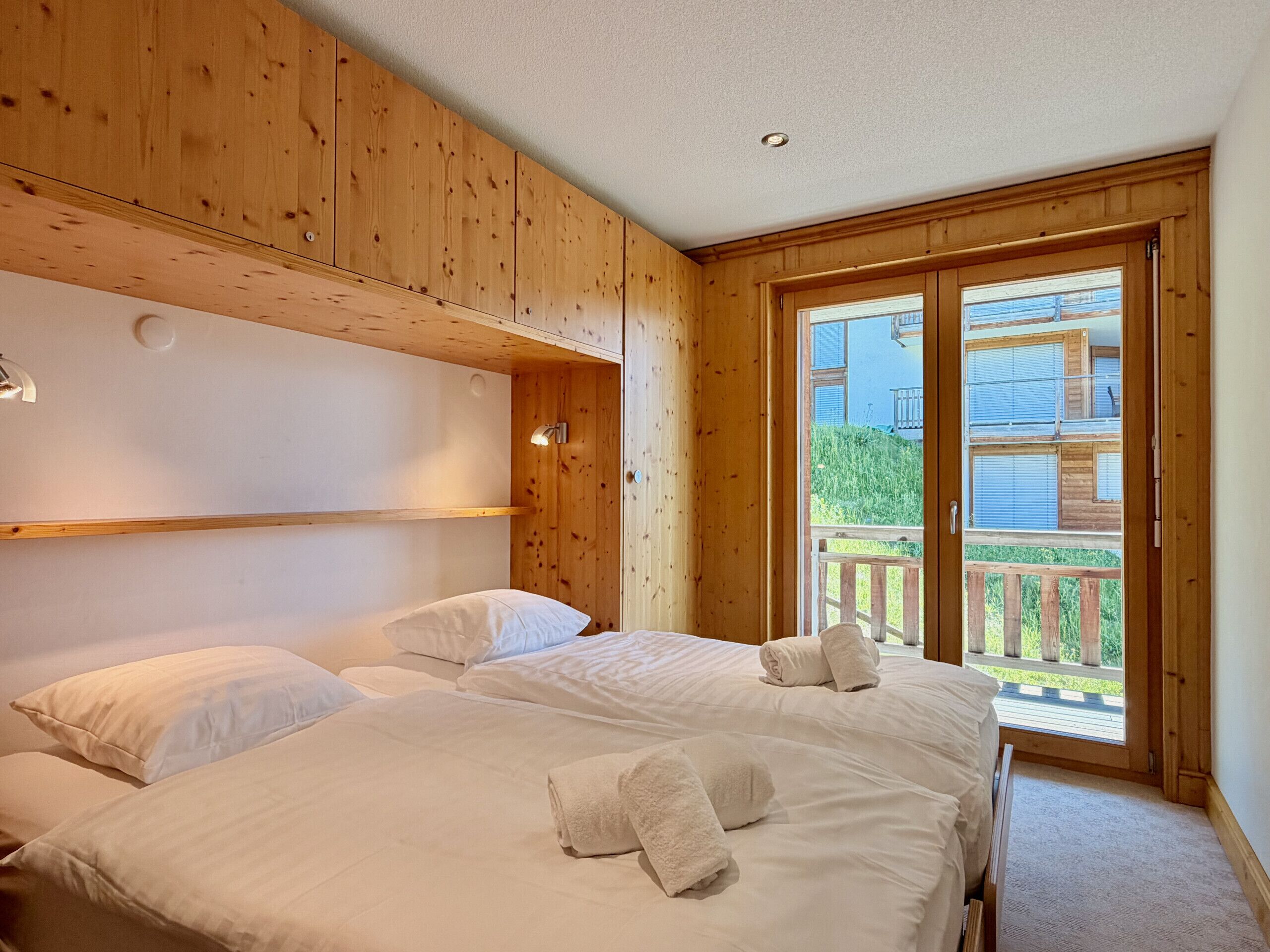 Ein holzverkleideter Raum im Blockhausstil mit zwei Betten, einem Balkon und Blick auf ein Gebäude.