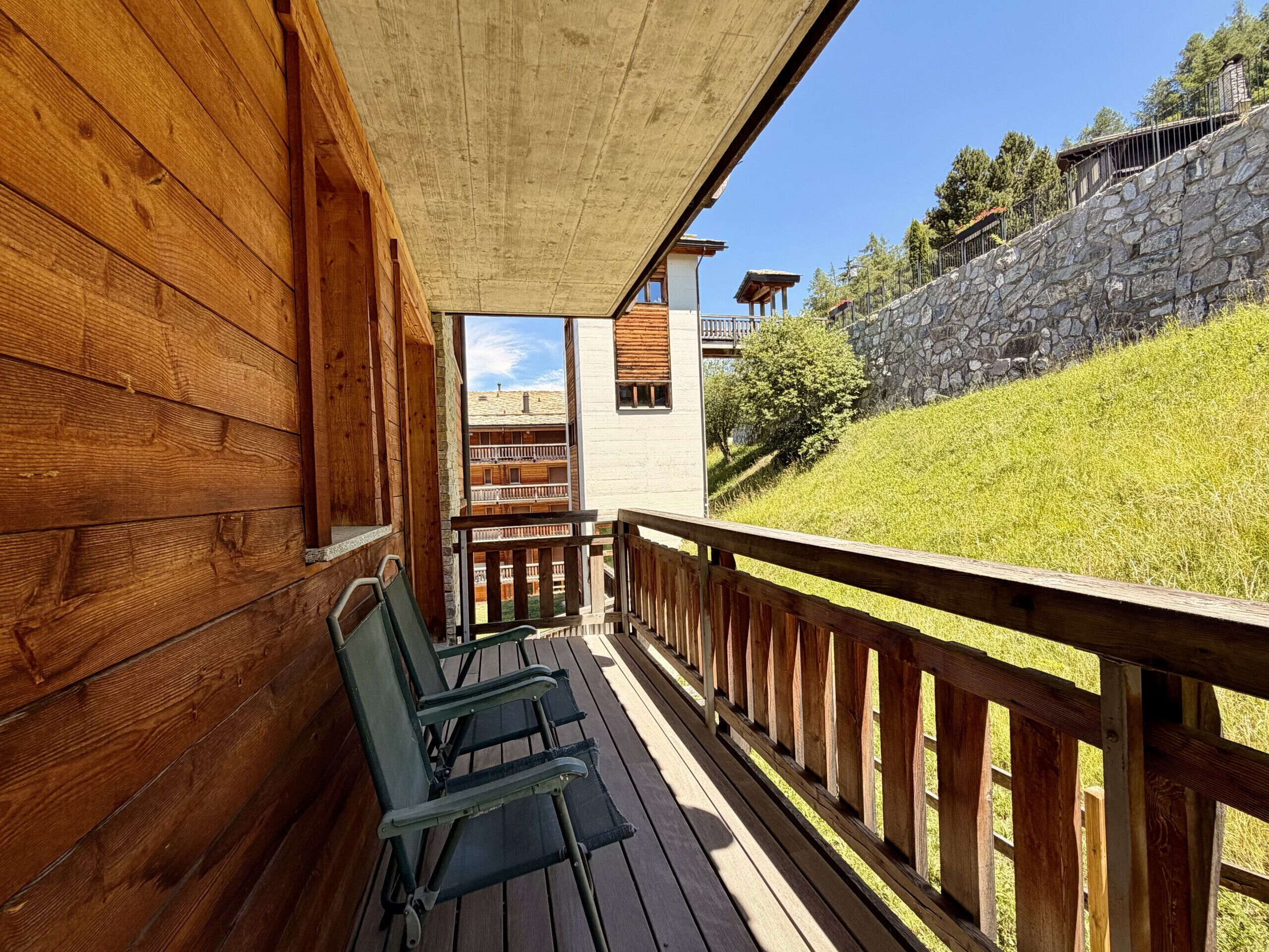 A wooden deck with chairs, a stone wall, and a view of a building with balconies.