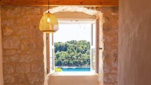 Un dormitorio con pared de piedra, techo de madera, cama con colcha blanca, mesita de noche con un libro y vista al mar azul a través de una ventana.