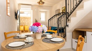 Un comedor con una mesa redonda de madera dispuesta para cuatro personas, un jarrón con flores y una escalera con barandilla de hierro forjado.