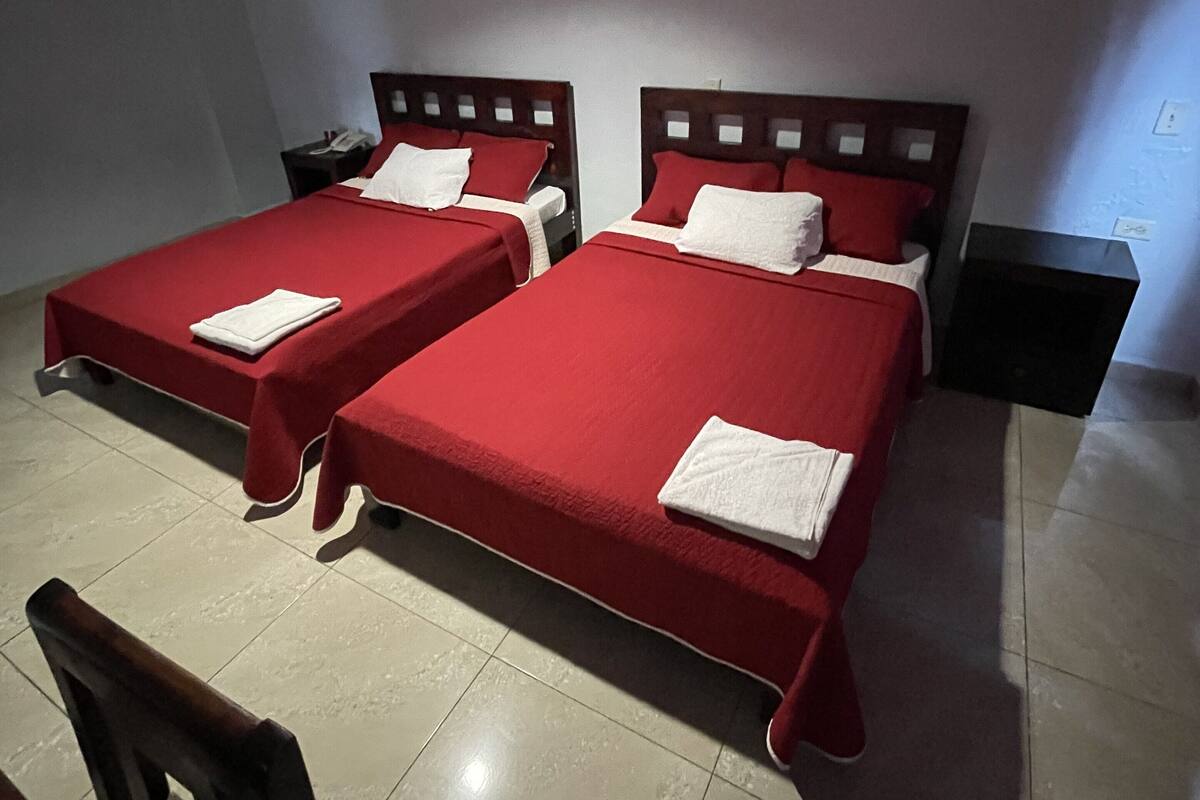 Une chambre d’hôtel avec deux lits simples, chacun recouvert d’une couverture rouge et agrémenté d’oreillers blancs. On y trouve une petite table avec une chaise, une table de chevet et une étagère fixée au mur.