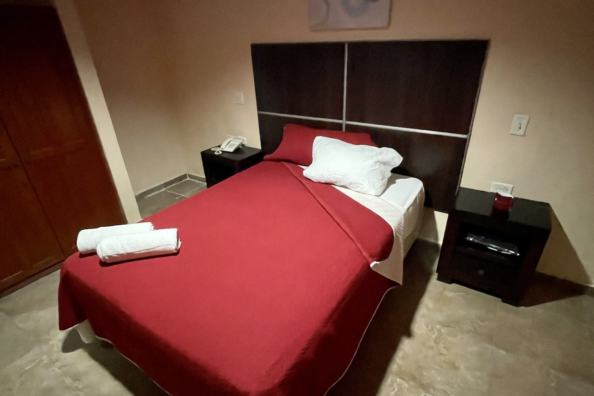 Un lit bien fait, avec une couvre-lit rouge et des oreillers blancs, une table de chevet avec un téléphone, et une œuvre d’art accrochée au mur au-dessus du lit.