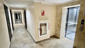 Een lange, smalle gang met betegelde vloer, een brandblusser bevestigd aan de muur, en een liftdeur in de verte.