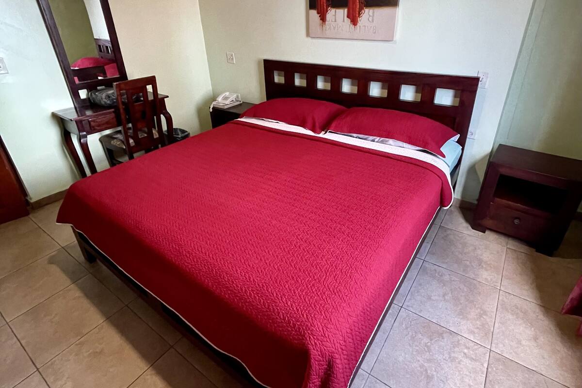 Une chambre avec un grand lit, une tête de lit en bois, une table de chevet, une chaise, un bureau et un tableau accroché au mur.