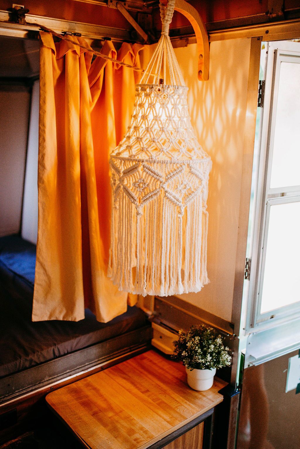 Un luminaire suspendu en macramé dans un intérieur chaleureux, agrémenté de rideaux et d’une petite table sur laquelle est posée une plante en pot.