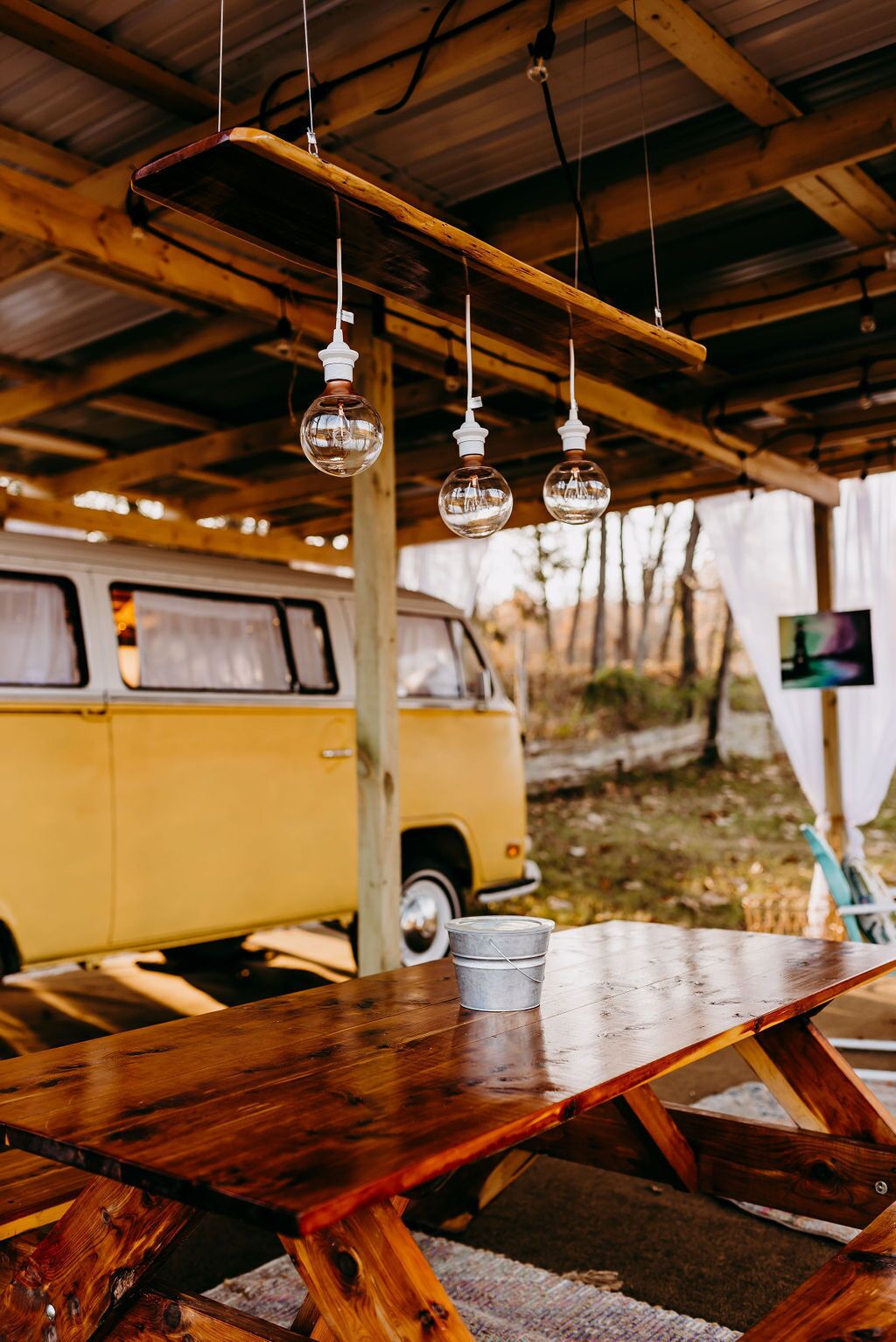 Une table en bois rustique, agrémentée de luminaires en verre suspendus, et un fourgon jaune en arrière-plan.