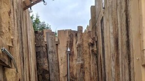 Una construcción rústica de madera con una estructura metálica en el centro, posiblemente un pozo o fuente de agua, sobre un suelo de grava.