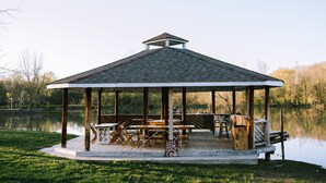 Een prieel met dak, houten tafels en stoelen, omgeven door een gazon en een waterpartij.