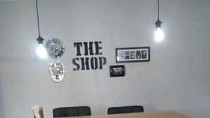 A dining area with a wooden table and four black chairs with chrome bases. There are two pendant lights hanging above the table. The wall behind the table has a sign that reads "THE SHOP" and a framed picture. - "THE SHOP" retro 60's (Keosauqua)