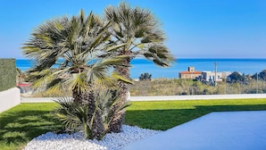 A modern outdoor area with a palm tree, white pebbles, and a view of the sea and buildings. - VILLA DARDA (Alcamo)
