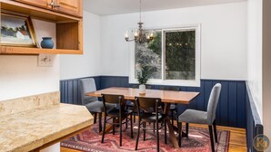 Un coin repas avec une table en bois, des chaises, un lustre, une fenêtre donnant sur des arbres et une étagère avec un vase et un tableau encadré.