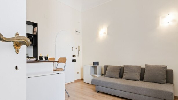 A modern living room with a sofa, a white cabinet, a door with a golden handle, and a wall-mounted light fixture. - GuestHost - Modern and cozy 25 sqm studio apartment, ideal for two people, located on the 2nd floor of a characteristic period building without lift. The property is located within the Traffic-Restricted Area of the historic center and is the ideal solut (Firenze)