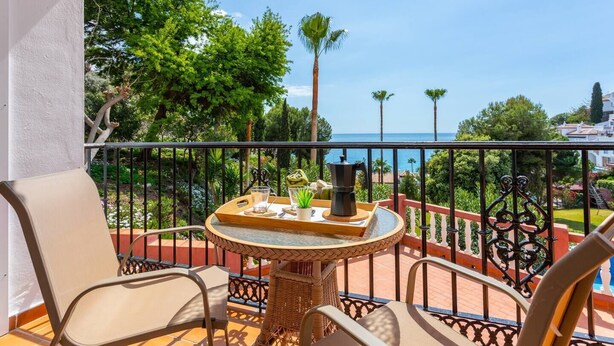 A balcony with wicker chairs, a round table with a tray of items, and a view of the sea and greenery. - Apartamento Ladera by Solset (Málaga)