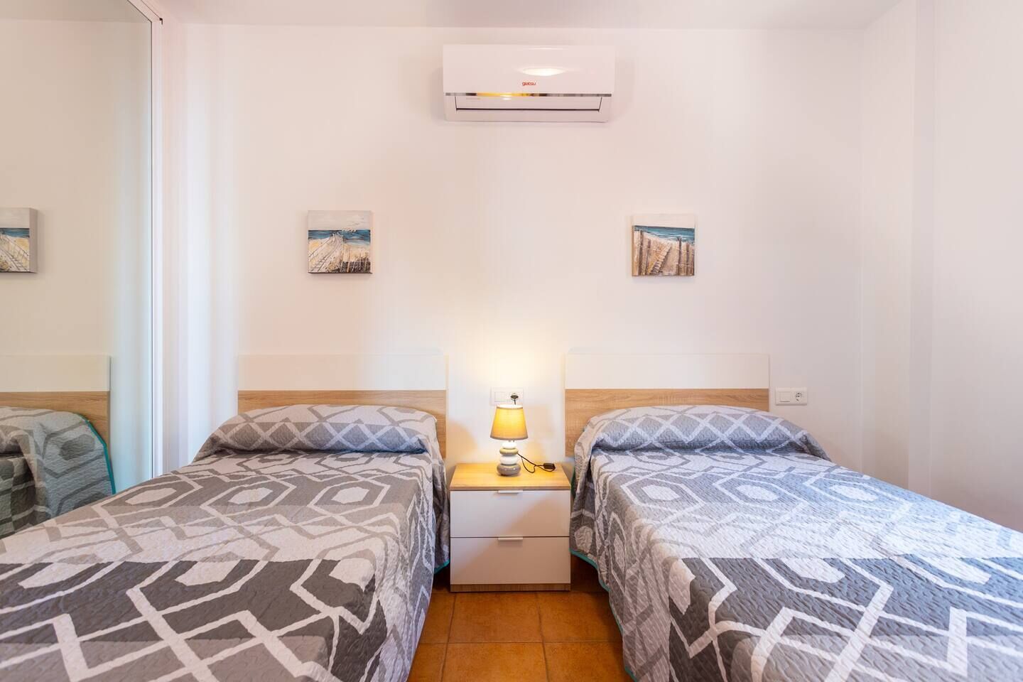 A hotel room with two beds, a nightstand with a lamp, an air conditioner, and two small paintings on the walls.