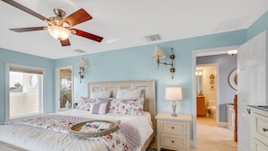 Une chambre à coucher avec un lit, des tables de chevet, un ventilateur de plafond et une salle de bain visible à travers une porte ouverte.