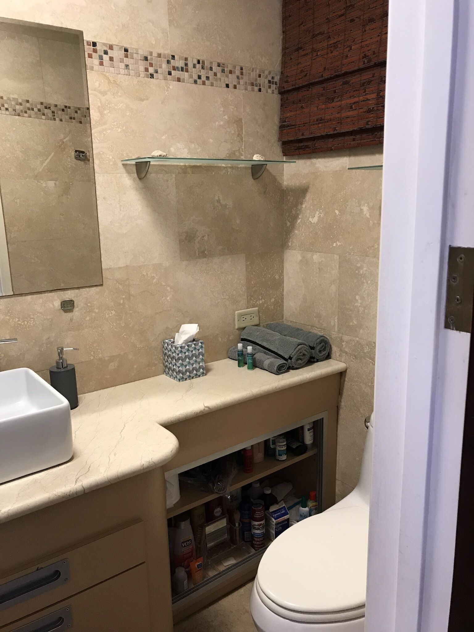 Une salle de bain avec un mur recouvert de carreaux beiges, une étagère en verre au-dessus du lavabo, des toilettes blanches et un meuble de rangement en dessous.